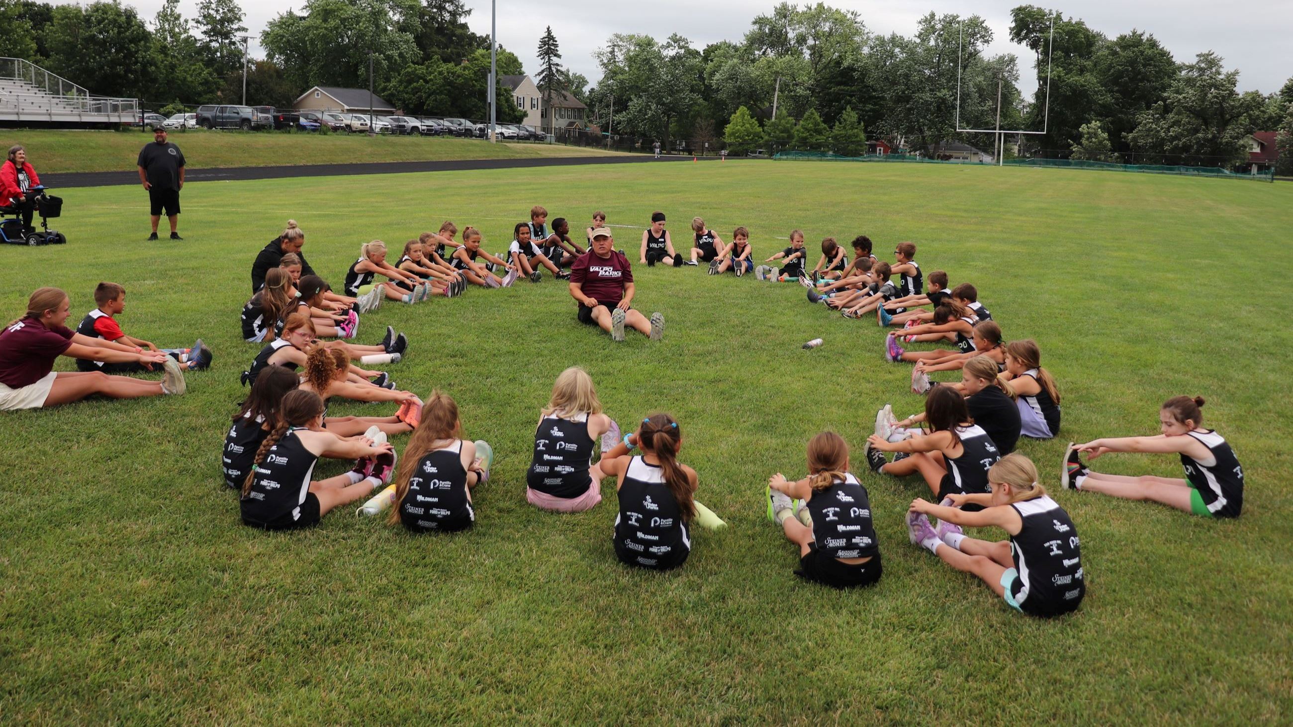 Track and Field participants in a circle around a coach, all stretching to touch their toes