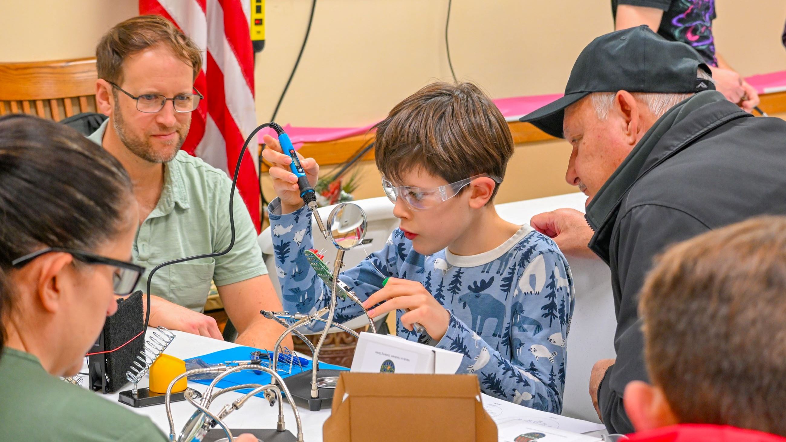 2 adults watching a boy with safety goggles working with soldering equipment