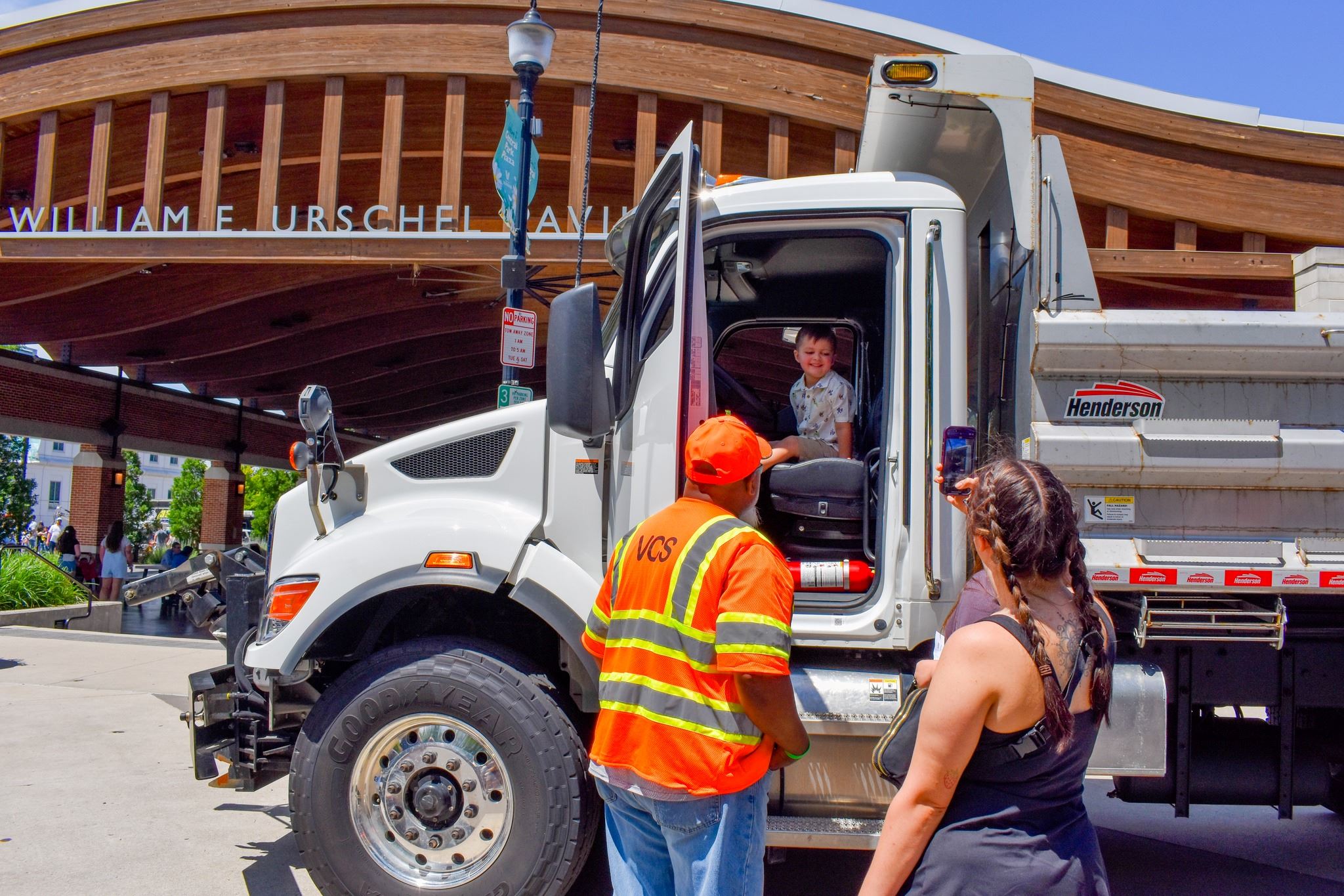 Child in drivers seat of a grey utility truck, with two adults outside the vehicle watching, one of which is taking a photo